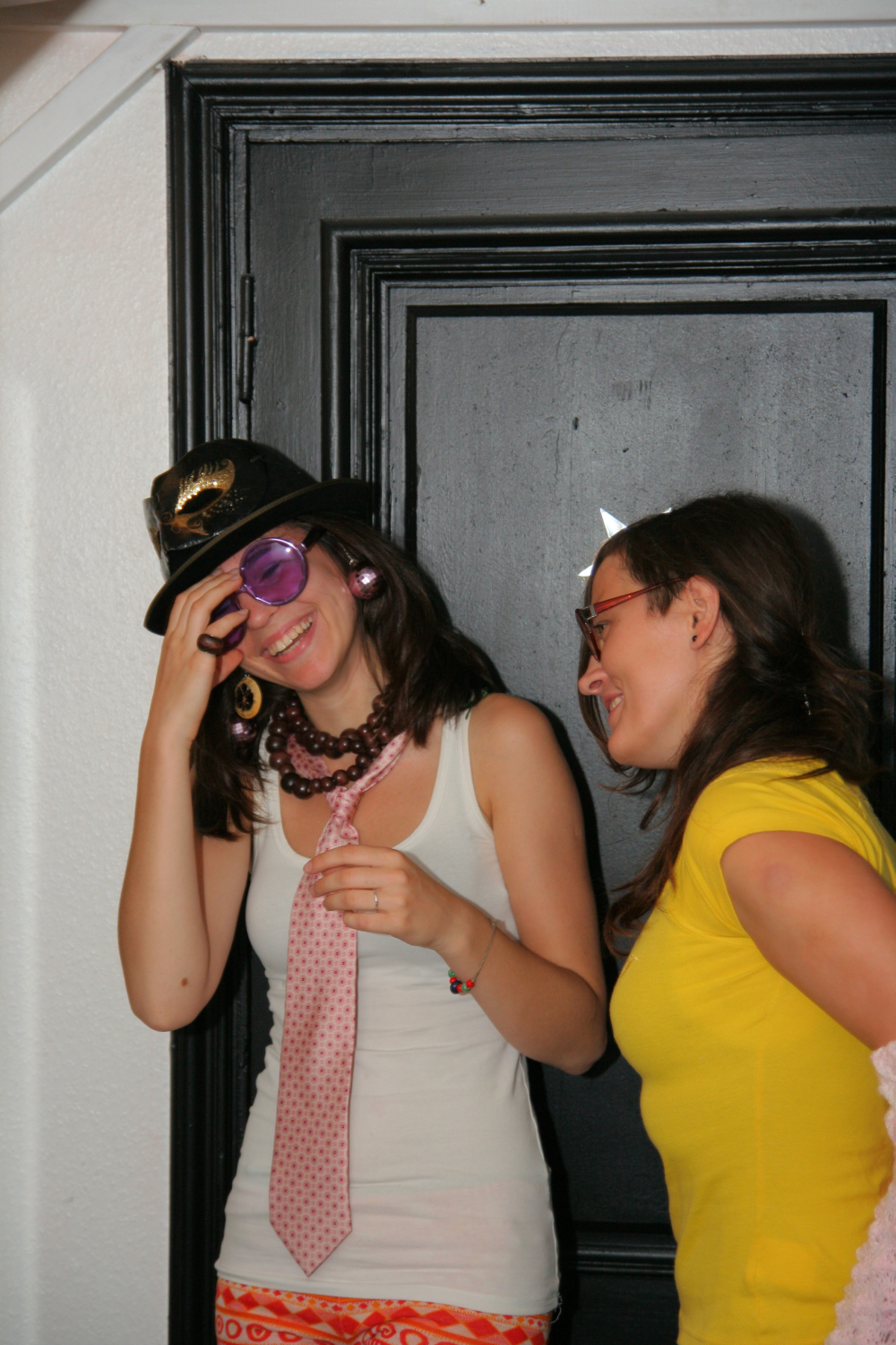 Two women are standing and smiling. The woman on the left is wearing a black hat, purple sunglasses, a white tank top, pink tie, and colorful pants. The woman on the right, sporting glasses with red horns and a yellow shirt, looks at her friend with amusement as they stand near a maiņas punkts against a black door.