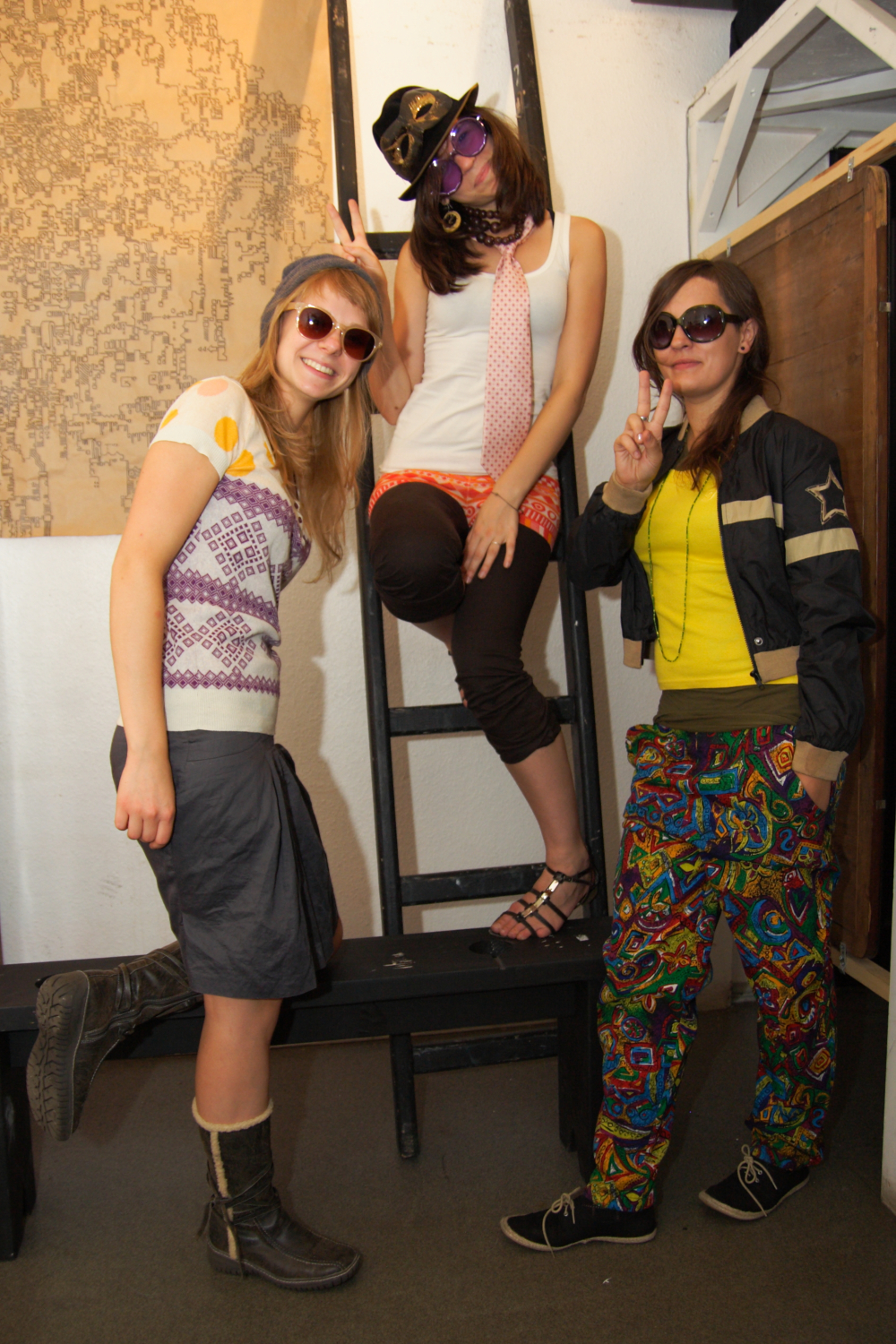 Three young adults wearing colorful and eclectic outfits pose cheerfully in an indoor setting, perhaps at a preparty. Two stand on the ground, while one sits on a ladder. They all wear sunglasses and make peace signs, exuding a fun, playful vibe. A wall map and wooden furniture are visible in the background.