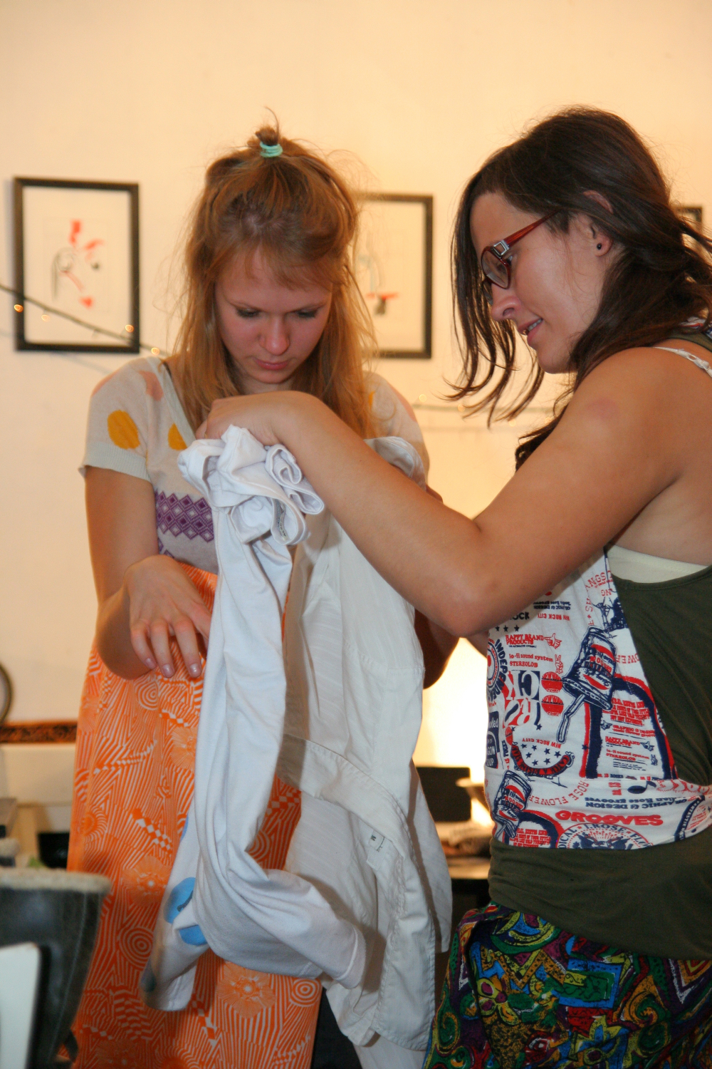 Two women are indoors, closely examining a white shirt. One woman with a colorful, sleeveless top and glasses is holding the shirt while the other woman in a patterned dress and a ponytail points to it. They seem focused and engaged, perhaps planning for their preparty. Several framed pictures hang on the wall in the background.
