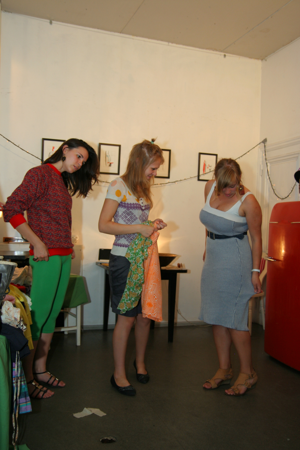 Three women are in a room decorated with framed pictures and string lights, clearly in preparty mode. One woman in a red sweater and green pants looks at her phone. The second woman, wearing a polka dot top, holds an orange-and-green fabric. The third woman in a striped dress looks down at her feet. A red appliance is seen on the right side at Maiņas punkts.