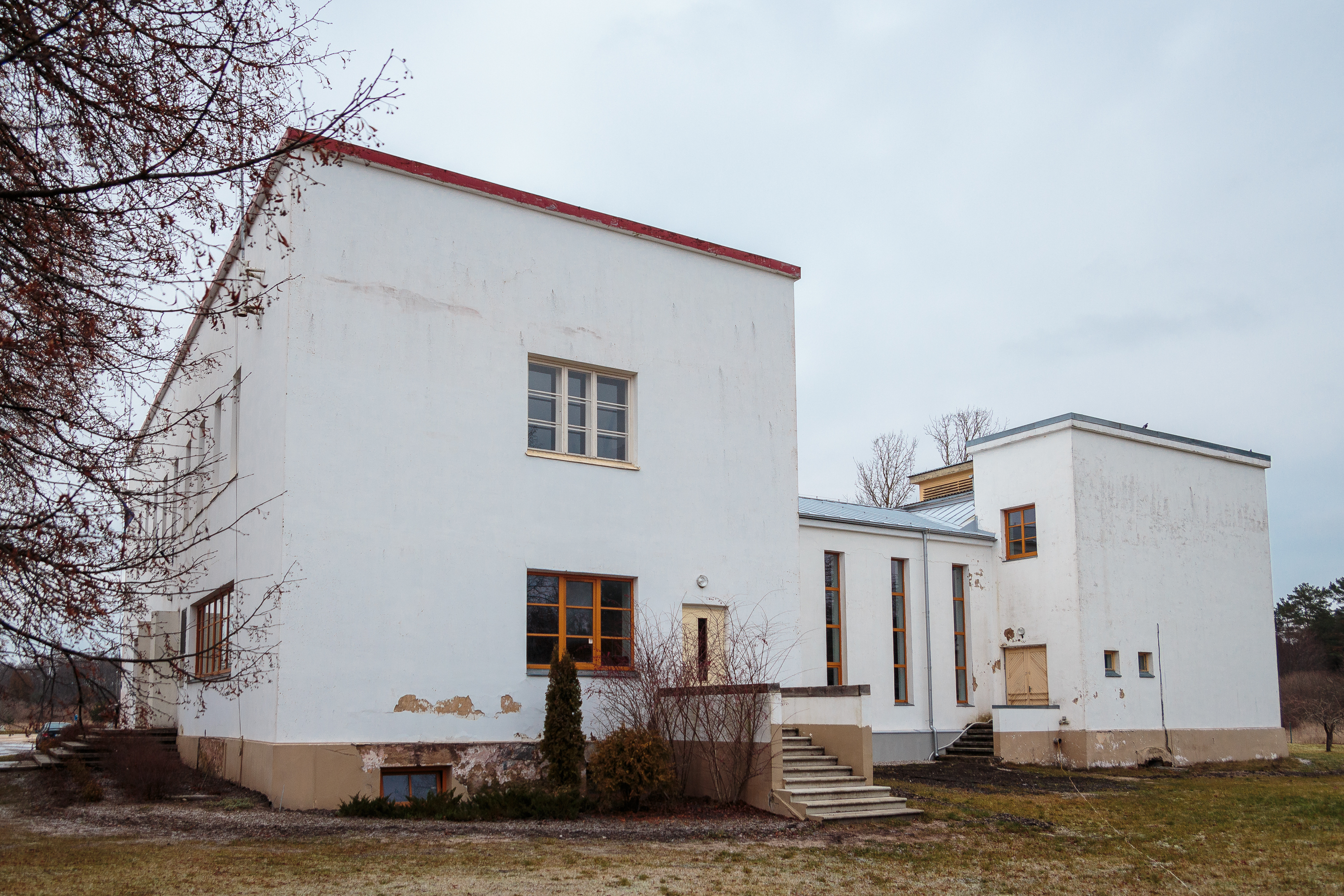 A two-story, Mazirbe-style white rectangular building with flat roofs and minimal architectural details features large windows and a small staircase leading to a door on the right. The exterior shows weathering and cracks, while sparse landscaping surrounds it under an overcast sky.