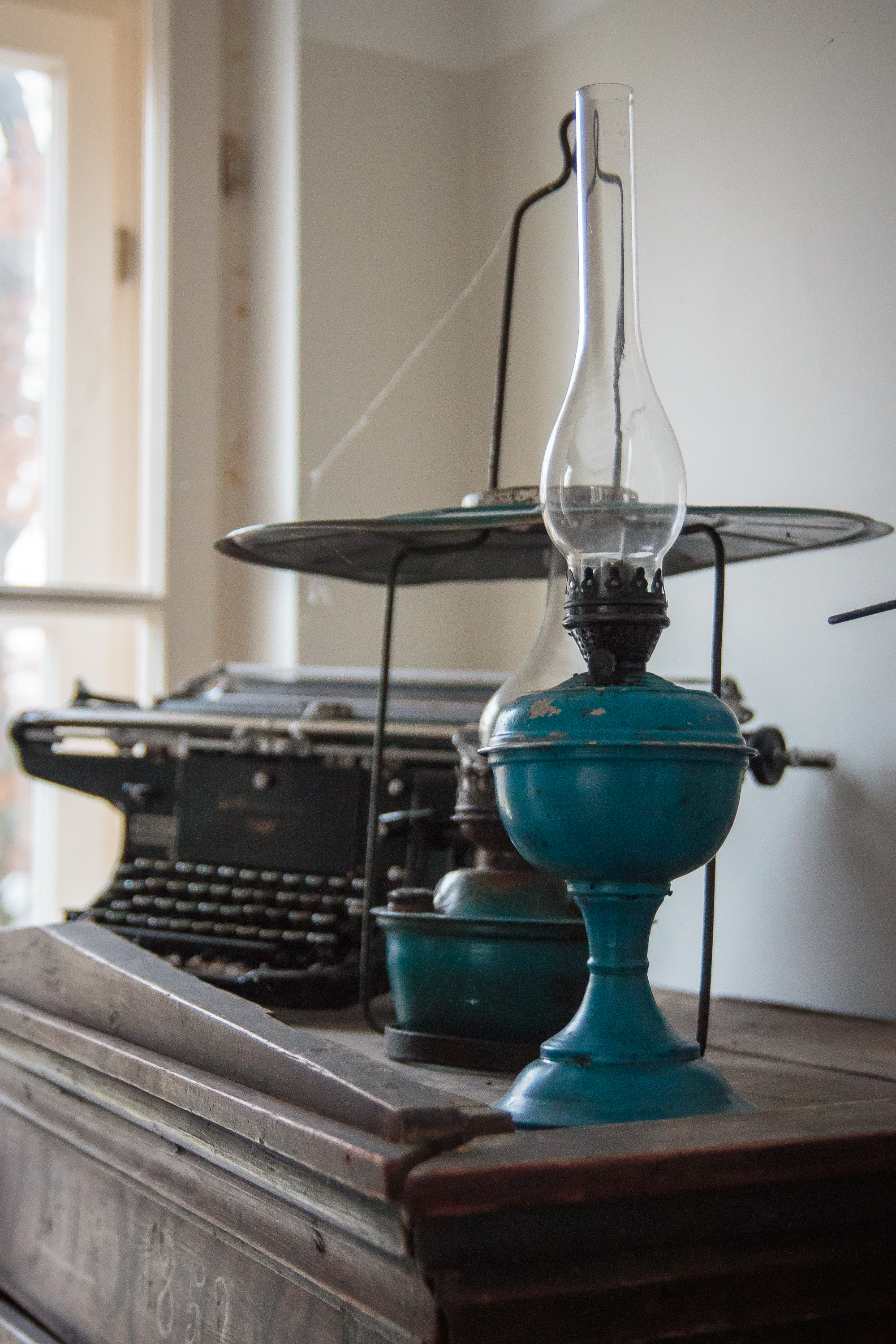 A vintage scene unfolds in Mazirbe, highlighting an old-fashioned typewriter and a blue oil lamp with a clear glass chimney atop a wooden chest. In the background, a balance scale by the window filters soft, natural light, evoking nostalgia and charm.