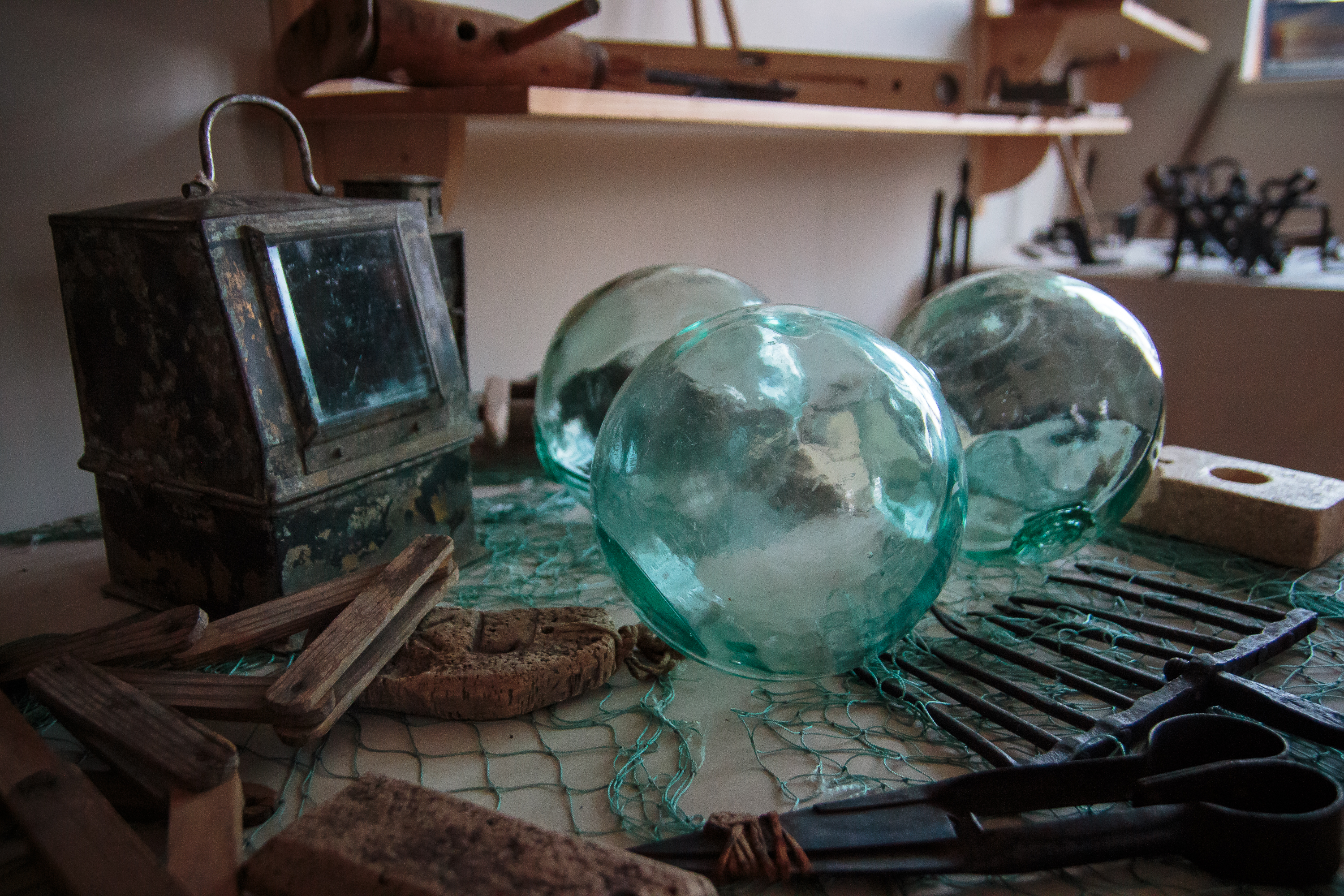 A rustic workbench in Mazirbe displays antique fishing gear: three large glass fishing floats, a weathered tin lantern, wooden net weights, and fishing nets. Assorted tools, including scissors, are scattered around, with shelves in the background holding more vintage items.