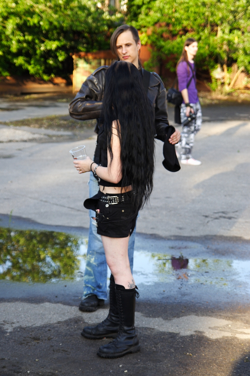 A person with long black hair, dressed in black shorts, a black top, and tall black boots, stands facing another person in a brown jacket and blue jeans. The scene resembles a moment from a 2009 metalshow. In the background, another person walks amid greenery while a puddle glistens on the pavement near their feet.
