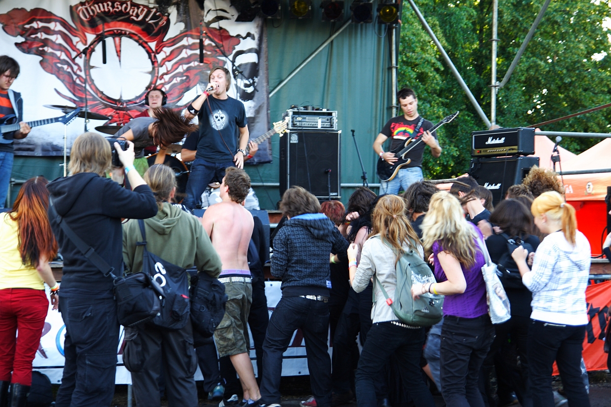A crowd at an outdoor metalshow enthusiastically watches a rock band perform on stage. The lead singer, dressed in a black shirt, is energetically engaging with the audience. The guitarist in a colorful shirt plays near an amplifier. Some of the attendees, their dyed hair reminiscent of 2009 trends, appear excited and energetic. There is a band banner in the background and trees are visible.