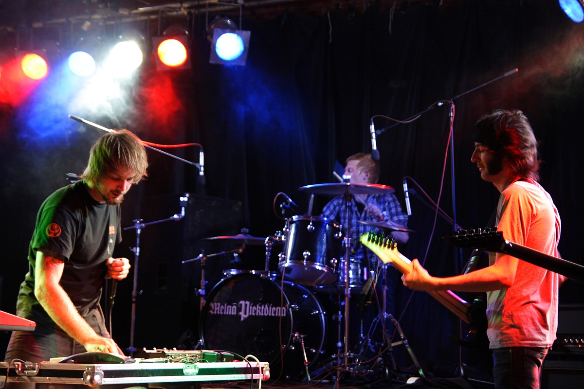 Three musicians perform on stage under colorful lights. The focus is on a guitarist wearing a dark shirt on the left, adjusting equipment at this 2009 metalshow. In the background, a drummer plays a drum set, and on the right, another guitarist in a light shirt faces the first guitarist. A dark backdrop completes the scene.