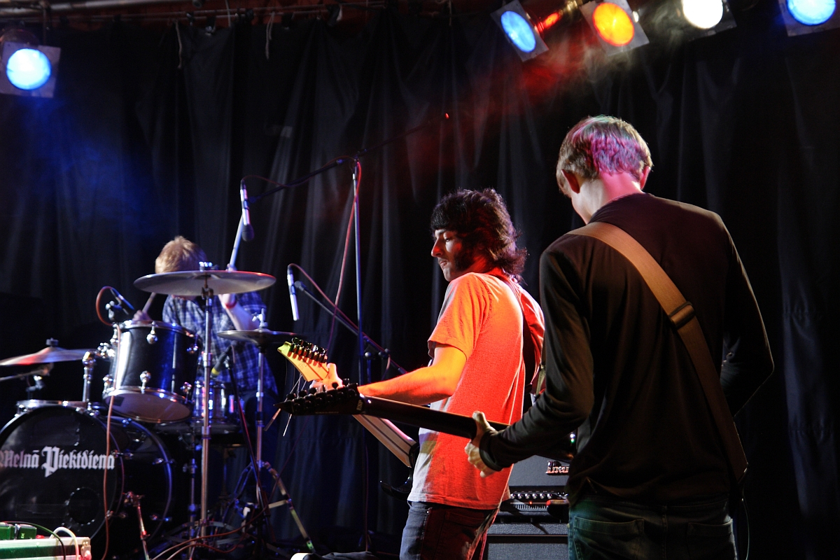 Three musicians are performing on stage. The drummer is playing a drum set, while two guitarists stand in front, one facing away and the other with long hair wearing glasses, looking down at his instrument. Colorful stage lights overhead illuminate the scene at Metalshow '09. The backdrop is black.