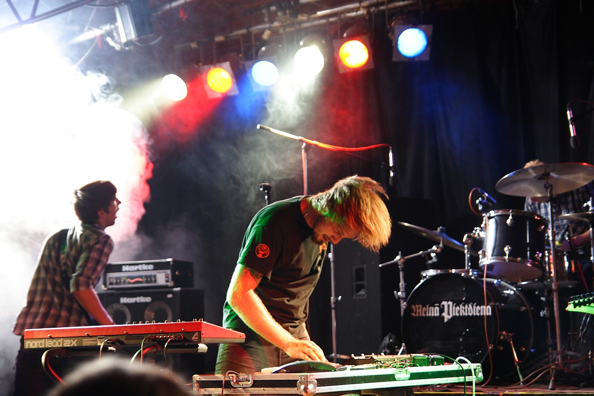 Two musicians perform energetically on stage at a 2009 metalshow, bathed in vibrant colored lights. One, wearing a black shirt, leans over an electric keyboard, hair swinging. The other, in a plaid shirt, is partially obscured. Smoke and stage equipment, including drums and amplifiers, fill the background.