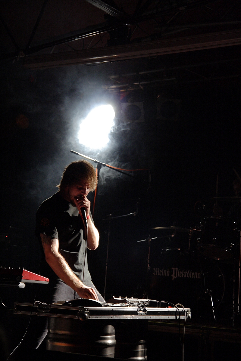 A person with medium-length hair performs on stage at a 2009 metal show, illuminated by a bright spotlight creating a smoky effect. They lean over a keyboard or synthesizer and hold a microphone close to their mouth. In the background, drumming equipment can be seen with "Mek Megtetem" written on the bass drum.
