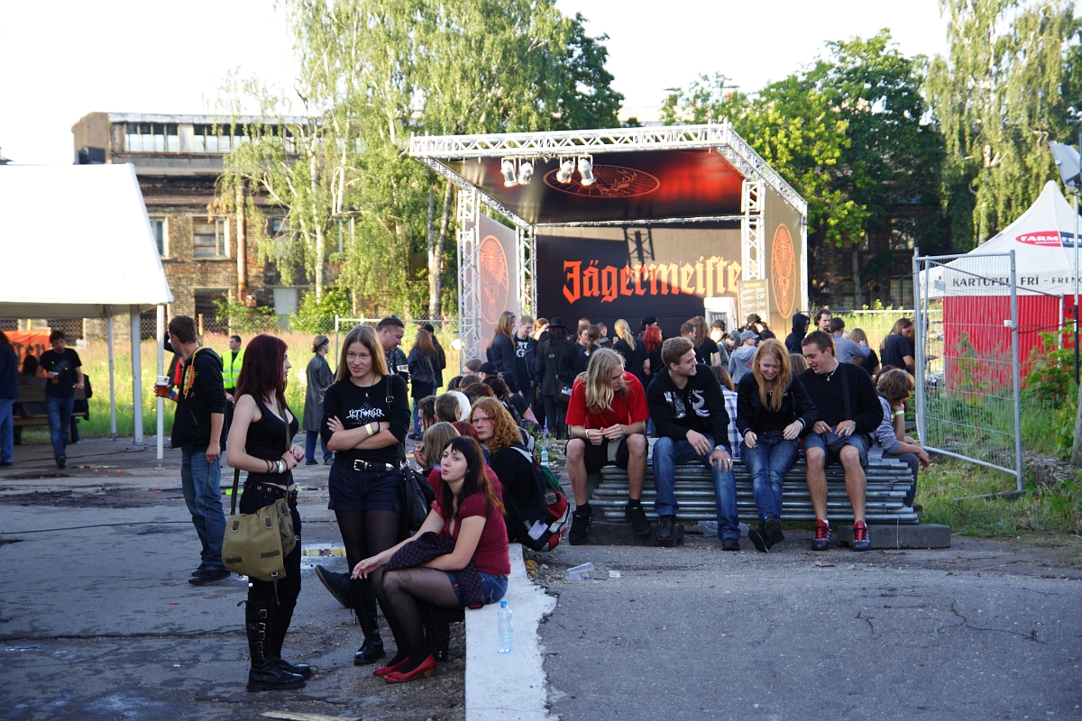 A group of people gather outside at a Jägermeister event. Some sit on steps in front of a stage with a Jägermeister banner, reminiscent of the Metalshow '09 atmosphere, while others stand or sit on the ground nearby. They appear to be socializing, and many are dressed in casual, dark-colored clothing. Trees and buildings are in the background.