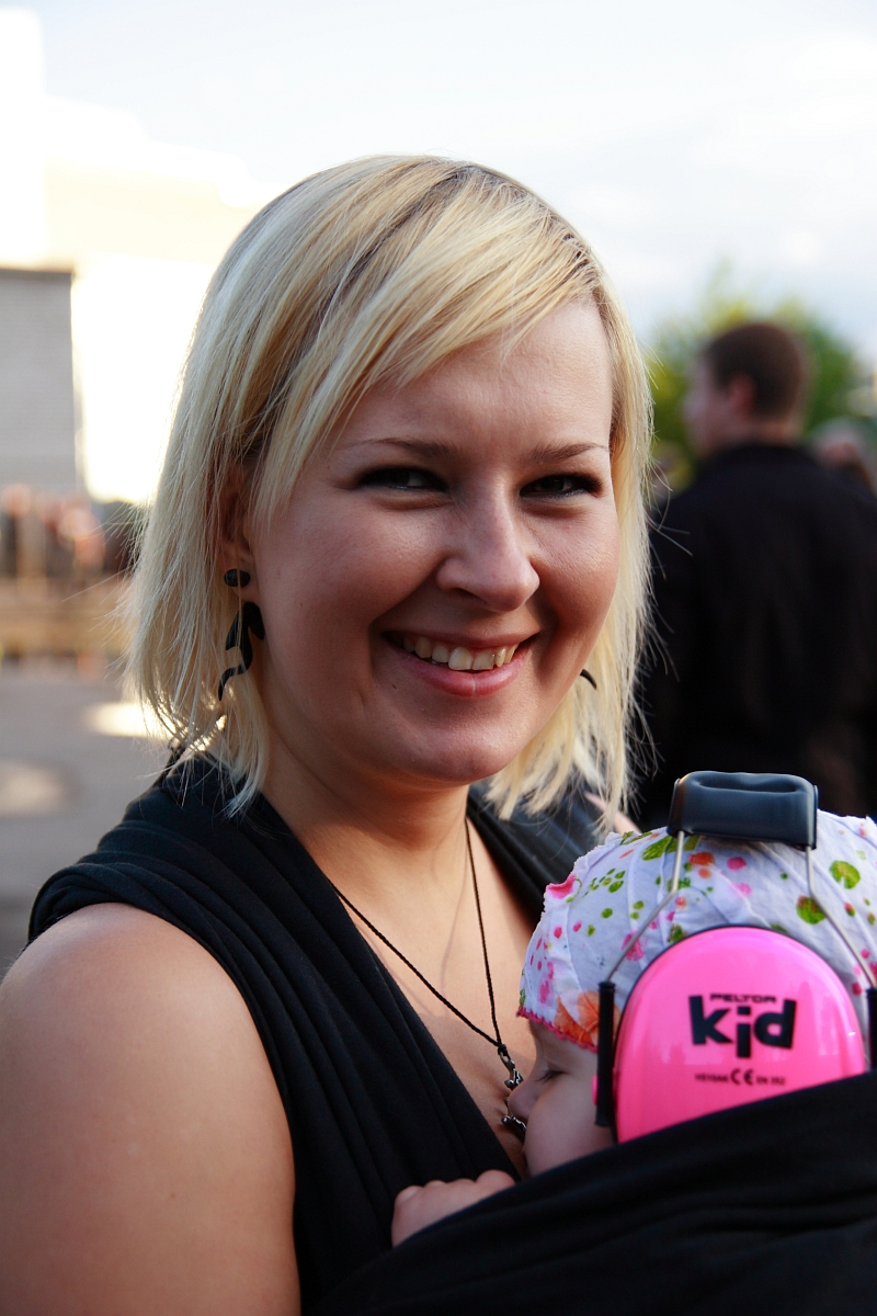 A smiling woman with short blonde hair is outdoors holding a baby, both enjoying what seems to be a lively Metalshow. The baby wears a white hat with colorful patterns and bright pink ear protection headphones labeled "kid '09." The woman, in black clothing and earrings, has blurred people and buildings in the background.