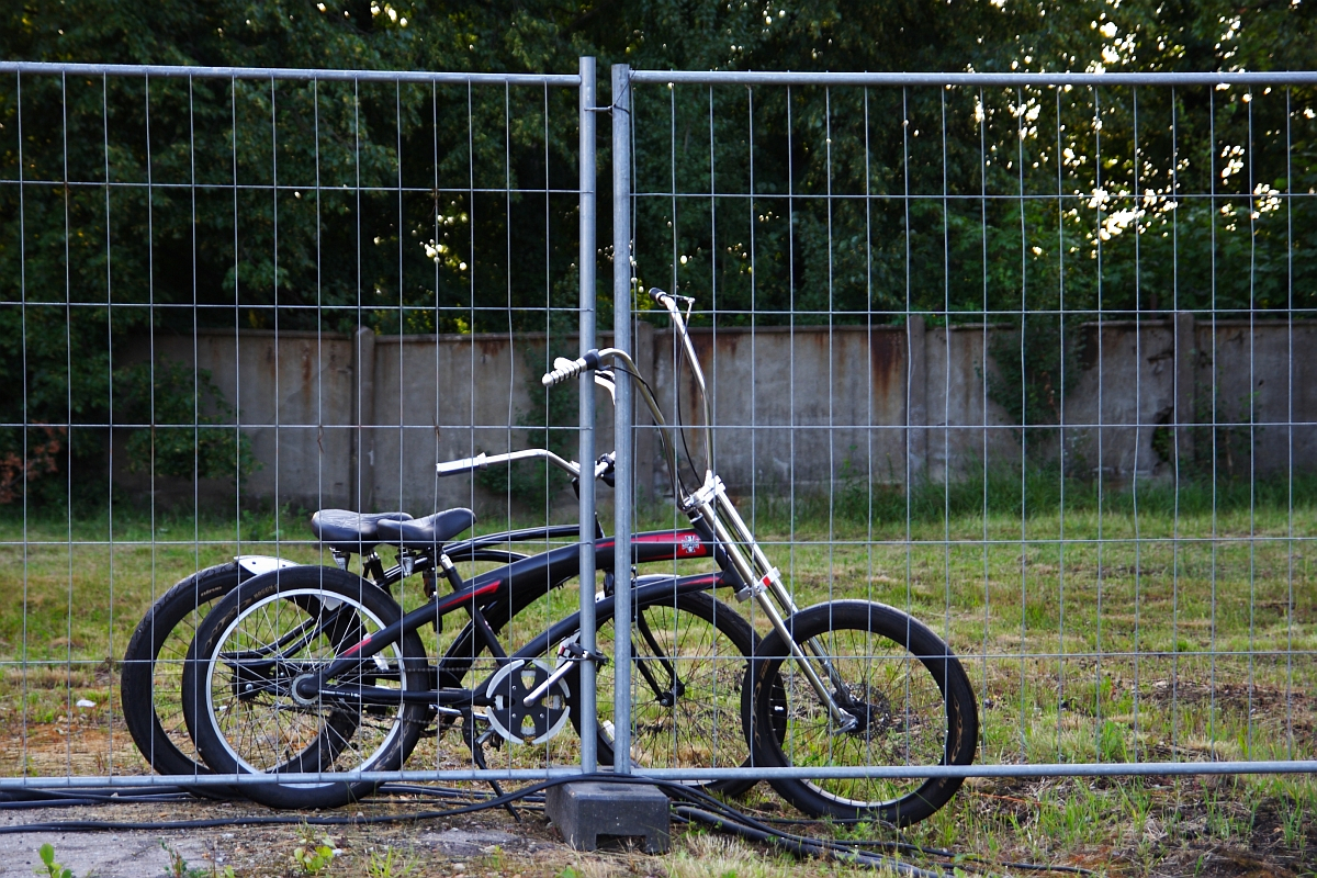 A black bicycle with a long front fork is locked to a wire mesh fence using a U-lock. The bike, reminiscent of the rides seen at Metalshow 2009, is parked on a grassy area with some exposed soil and scattered vegetation. Behind the fence, there is a concrete wall and a backdrop of trees in a wooded area.