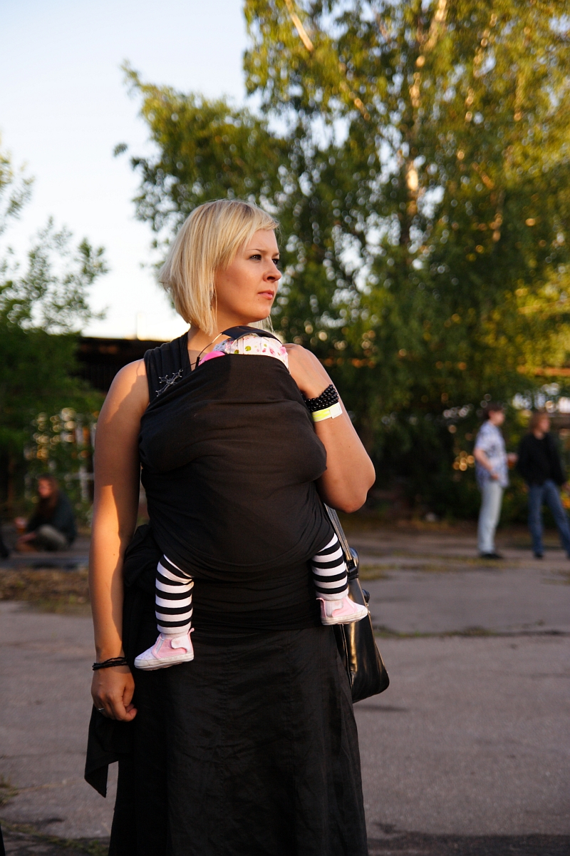 A woman with short blonde hair stands outside on a paved area with trees in the background, reminiscent of a scene from 2009. She wears a black dress and holds a baby in a black wrap carrier, their legs clad in black and white striped leggings and pink shoes protruding. Other people reminiscent of a metalshow are in the background.