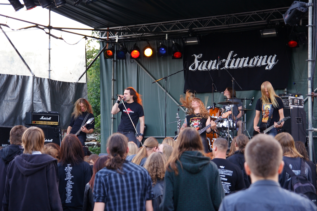 A band performs on an outdoor stage with a banner behind them reading "Sanctimony." The lead vocalist sings into a microphone, while other members play electric guitars and drums. The audience below, comprised of people with long hair and back-printed shirts, watches the 2009 metalshow.