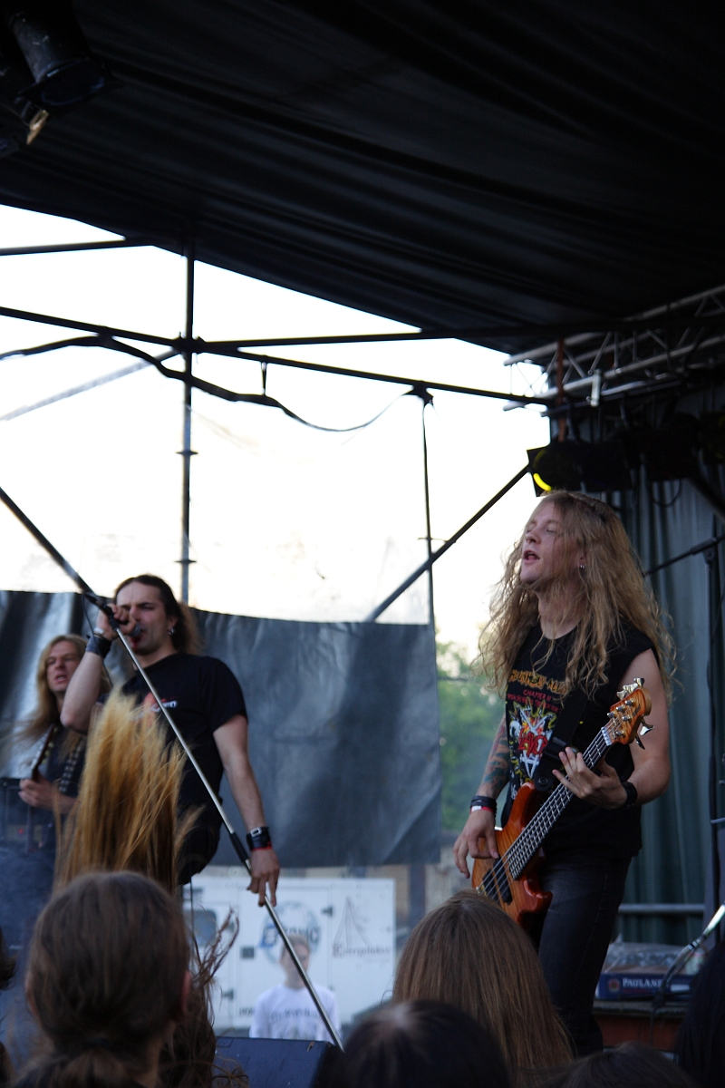 Two musicians performing on an outdoor stage at Metalshow '09. The one on the left is singing into a microphone, while the one on the right, with long blonde hair, is playing a bass guitar. Both are wearing black T-shirts, and the stage backdrop is dark with a partially visible drum kit. An audience with long hair is visible in the foreground.