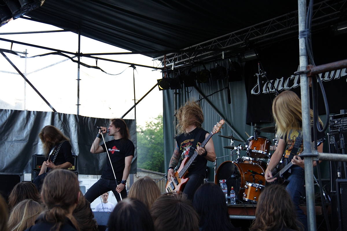 Band performing on outdoor stage at Metalshow '09. Five male members with long hair are playing instruments including guitars, bass, and drums. Lead singer is gripping a microphone stand. Audience with blond and brown hair visible in foreground. Backdrop features the word "Sanctuary.
