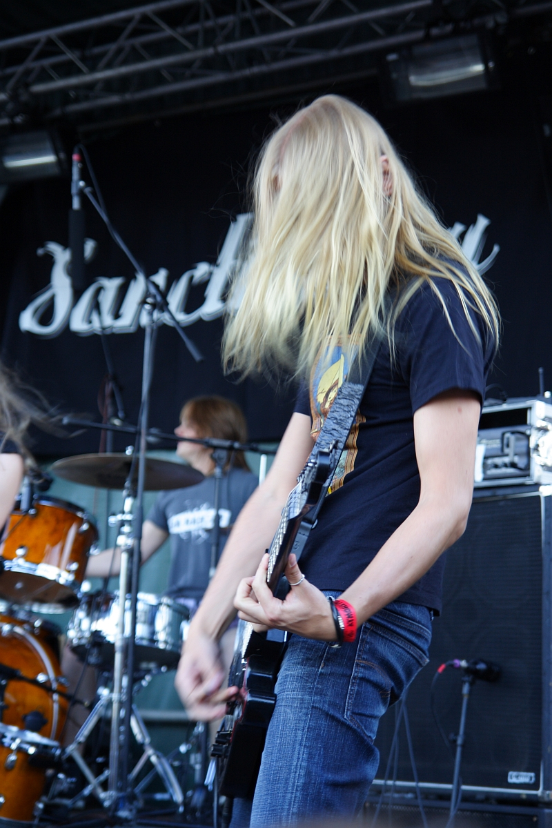 A person with long blonde hair is playing a guitar on stage at Metalshow '09, moving their head vigorously. They are wearing a black t-shirt and jeans. In the background, another person is playing drums. The backdrop features the partially visible word "Sanctuary" in stylized, white lettering.