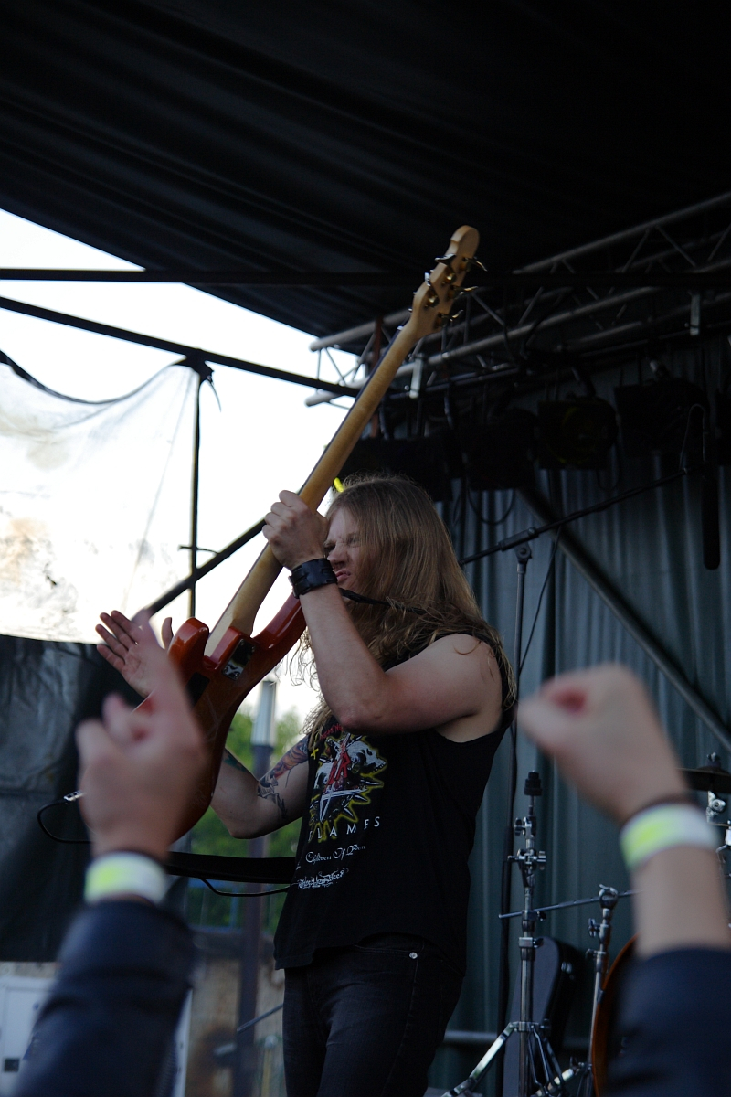 A long-haired musician in a black sleeveless shirt with a colorful design plays an orange electric guitar on an outdoor stage at Metalshow '09. He is captured mid-performance raising his arm. The audience, with raised arms and wristbands, is visible in the foreground. Stage lights and equipment are in the background.