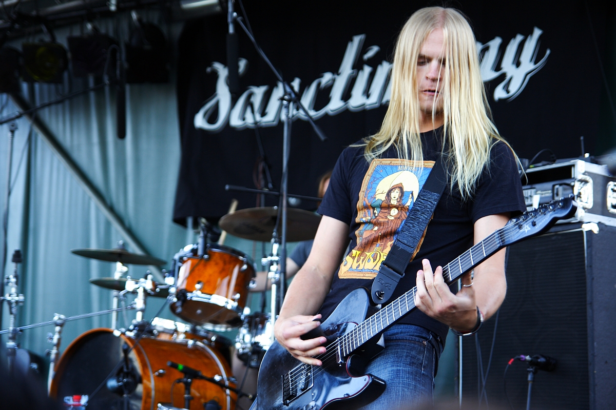 A long-haired musician plays an electric guitar on an outdoor stage at a 2009 metalshow. He wears a black T-shirt with a colorful design. Behind him are a drum set and other musical equipment. A black banner with the word "Sanctuary" is hung in the background.