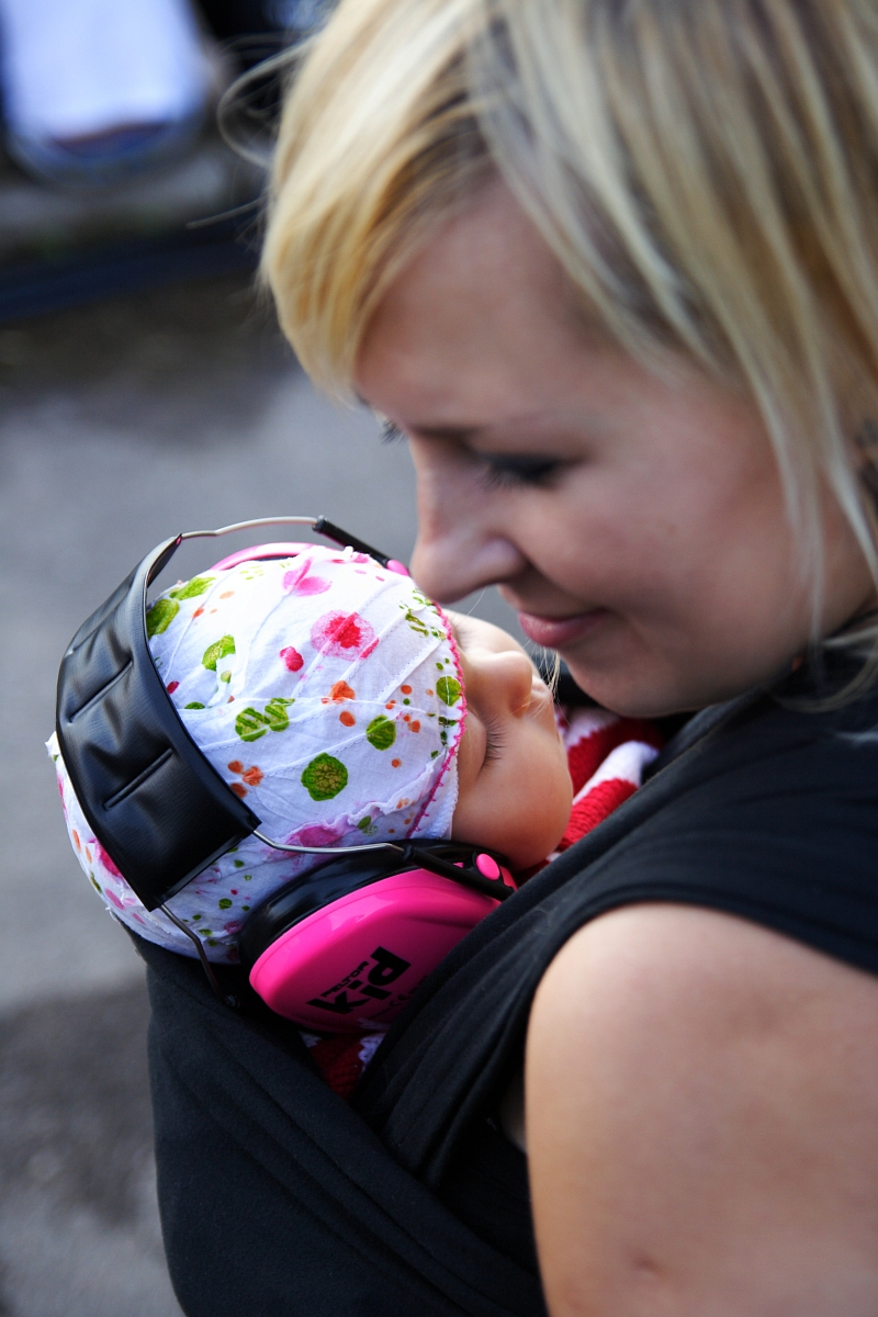 A woman with blonde hair is carrying a baby with floral-patterned headwear in a baby carrier at Metalshow '09. The baby, wearing pink ear protection headphones, appears to be asleep. The woman is smiling and looking down fondly at the baby while the background is out of focus.