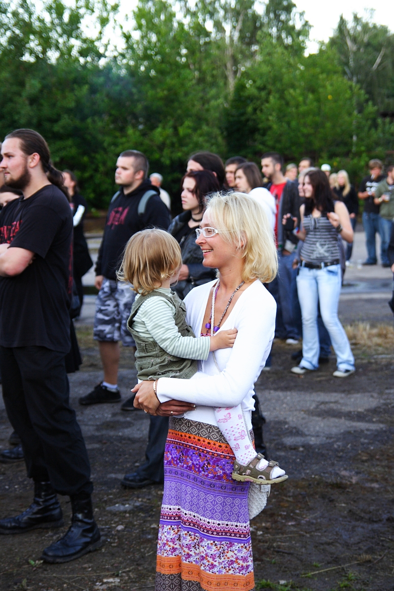 A woman with blonde hair, sunglasses, and a colorful skirt holds a young child in her arms at Metalshow 2009. The child wears a striped shirt and green vest. They are surrounded by other attendees, some in dark clothing, with greenery in the background. Several people observe something off-frame.