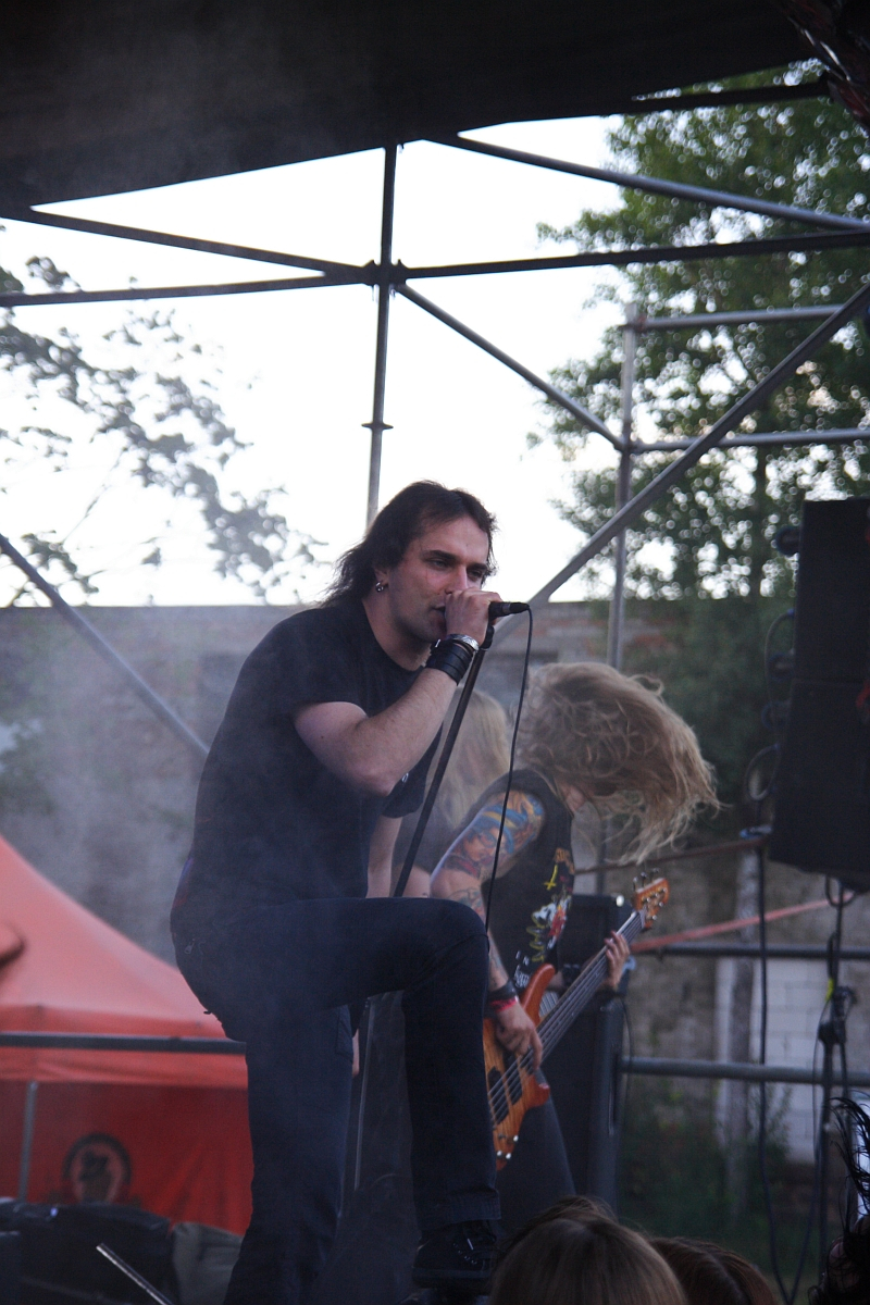 A lead singer on stage passionately sings into a microphone while a guitarist with long hair headbangs in the background at this 2009 metalshow. The stage features metal scaffolding and a red canopy. Trees and some blurred audience members are visible in the background, adding to the outdoor concert ambiance.
