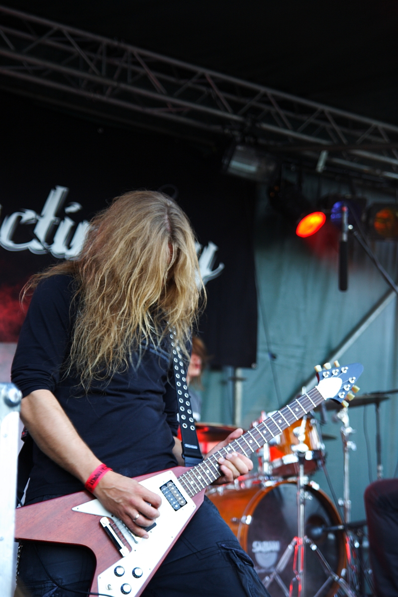 At a 2009 Metalshow, a musician with long blond hair covering their face plays an electric guitar on stage. They are wearing a black shirt and have a red wristband. The guitar has a distinct V shape with a white body and dark edges. Behind them, a drummer plays under red and orange stage lights.