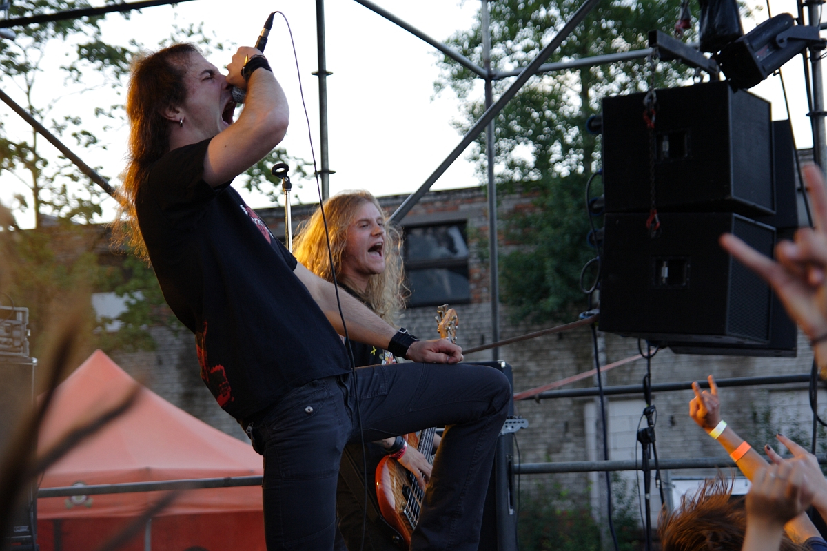 Two musicians perform passionately on an outdoor stage at the 2009 metalshow event. The lead singer, with long hair and a black t-shirt, leans forward while gripping the microphone. The bassist, also with long hair, plays intensely beside him. Fans' hands and stage equipment are visible, with a brick building in the background.