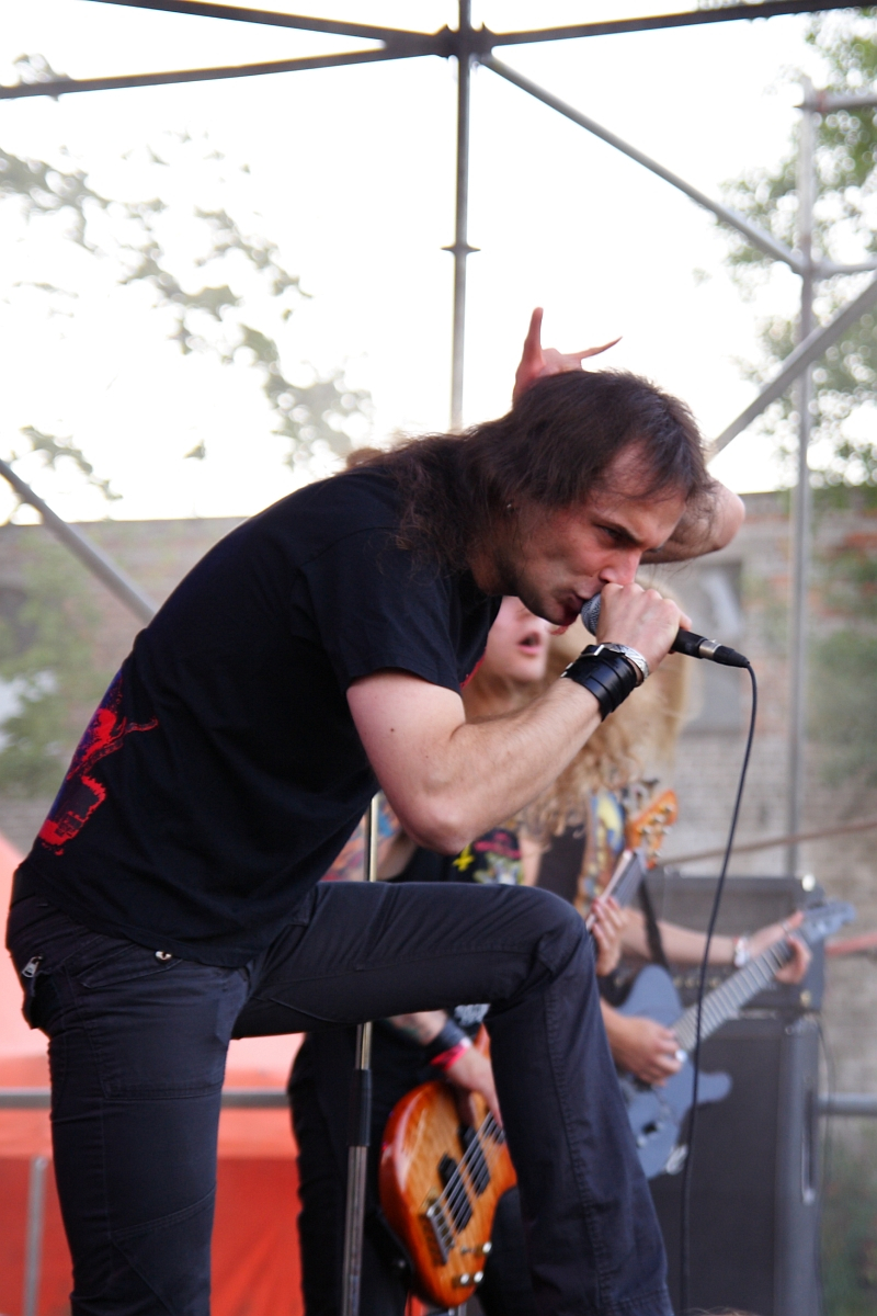 A man wearing black clothing, including a black shirt and pants, is performing on stage at a 2009 Metalshow. He grips a microphone with his right hand, his left fist raised. A musician with long blonde hair and a bass guitar stands in the background. Metal scaffolding or stage equipment is visible around them.
