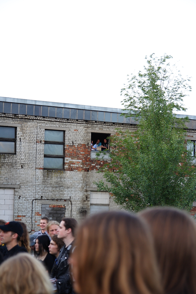 A crowd of people is gathered in an outdoor area near a worn-out two-story brick building. Several attendees of Metalshow '09 look out of a large window on the building's second floor. The building shows signs of decay with patches of missing bricks, while a tall tree stands in front.