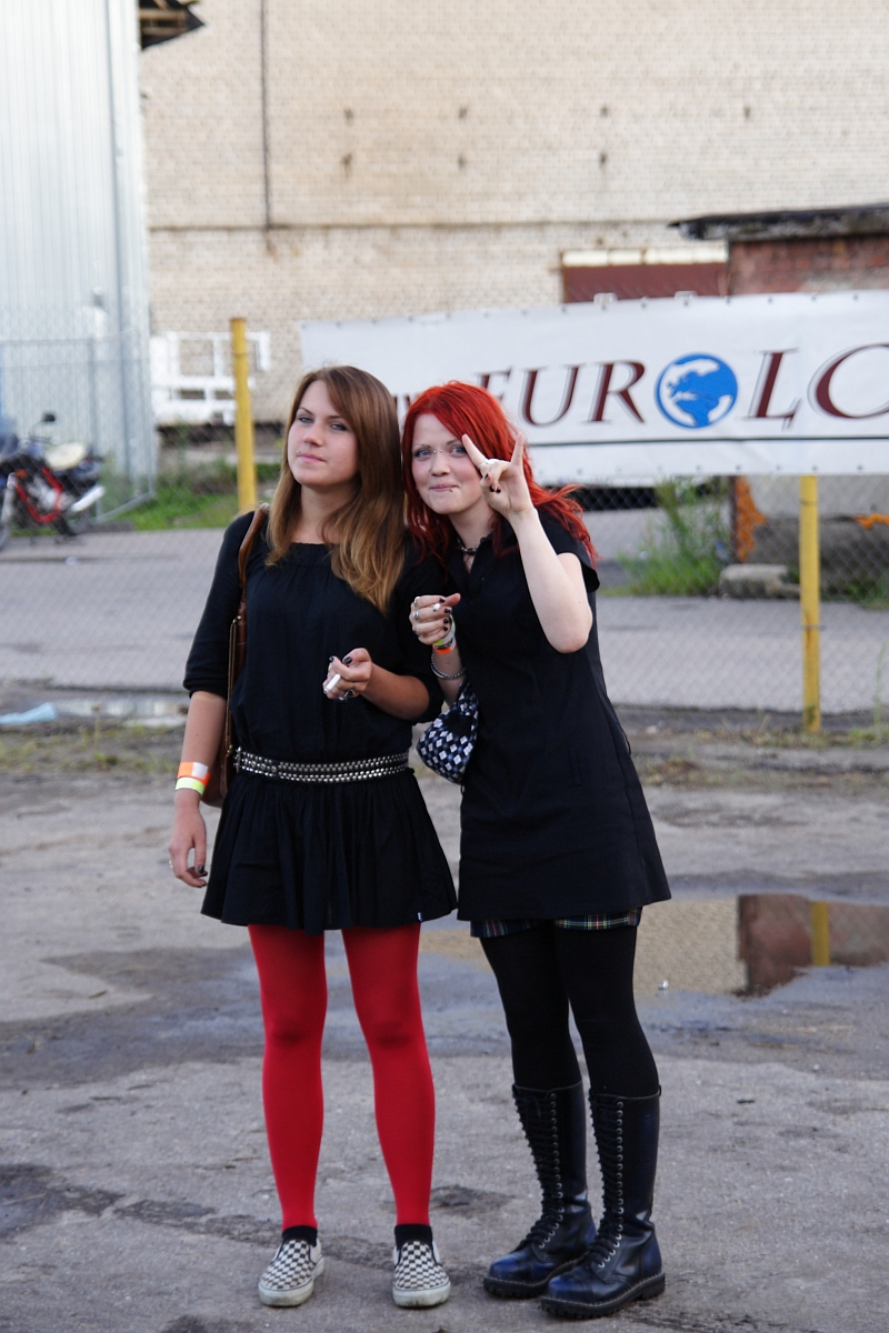 Two young women stand outdoors in front of a chain-link fence with a EuroLC sign, reminiscent of an industrial metalshow setting. One, with red hair and a black dress, makes a peace sign while holding ice cream. The other, with brown hair in a black dress and red tights, looks off to the side. It's 2009; bikes and industrial structures complete the background.