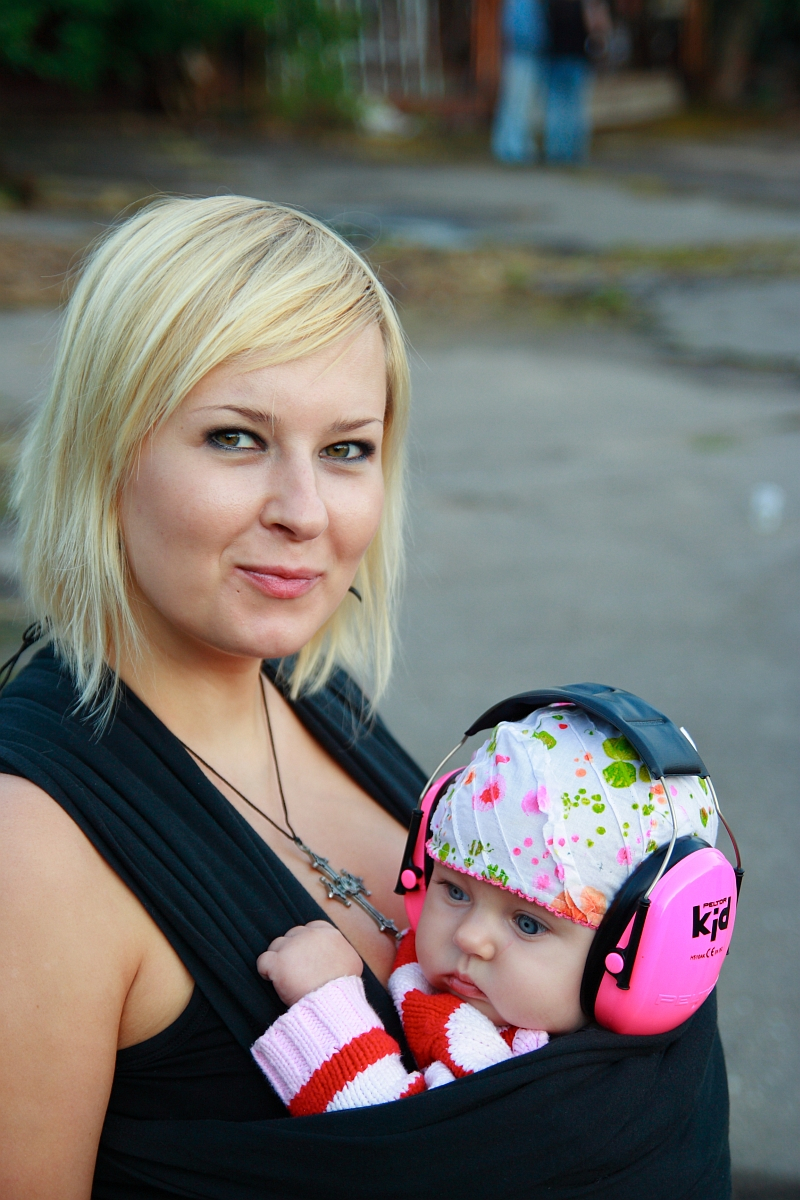 A blonde woman with a slight smile carries a baby in a black baby carrier. The baby is wearing a white hat with colorful patterns, pink earmuffs labeled "kid," and a striped sweater. They are outdoors at an event, possibly reminiscent of the bustling streets during Metalshow 2009, with a blurred urban background.