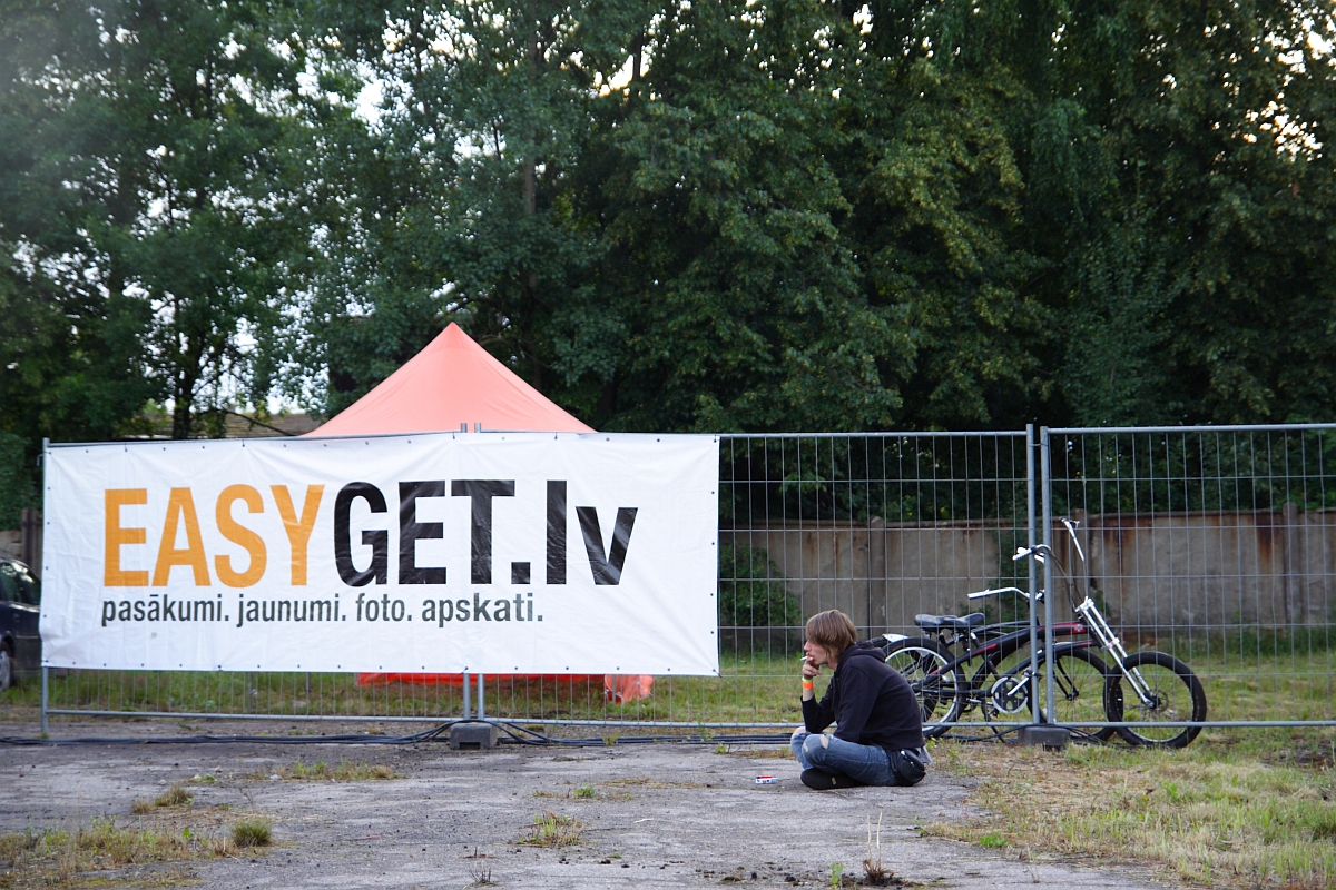A person sits on the ground outside, leaning against a metal fence. Behind the fence, a large white banner reads "EASYGET.lv pasākumi, jaunumi, foto, apskati." Nearby are two bicycles and an orange tent set up in front of dense green trees. It's clear this spot is ready for Metalshow '09.