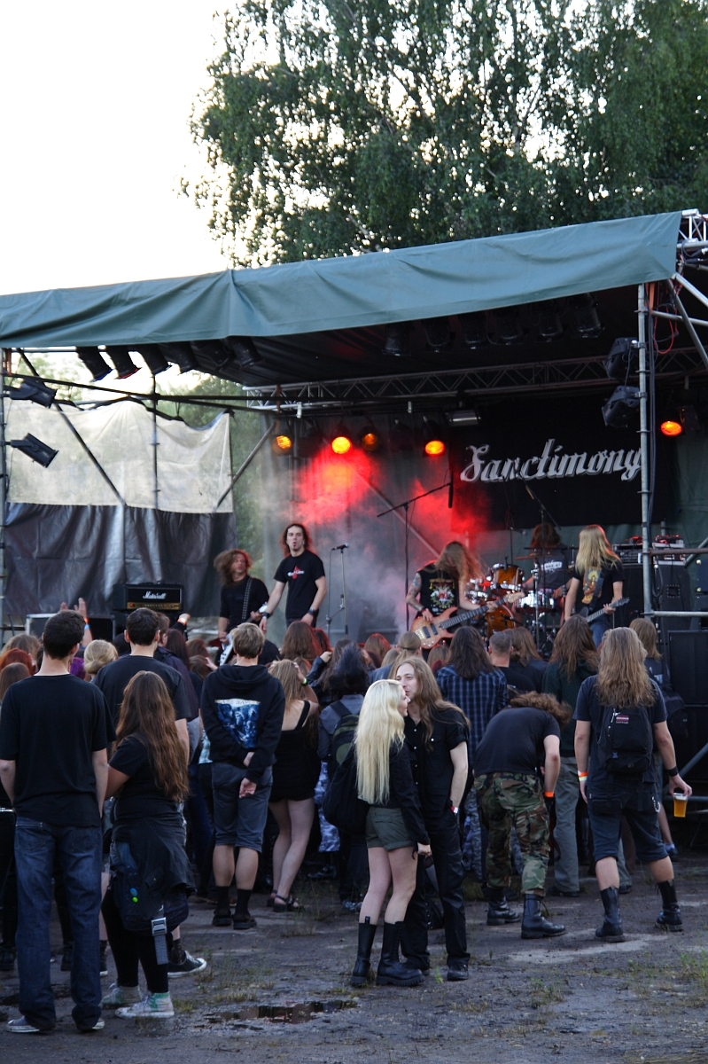 A crowd gathers in front of an outdoor stage where a metal band is performing at Metalshow '09. The band members are engaged with their instruments and singing under colorful stage lights. The audience, dressed in dark clothing, watches attentively. Trees and outdoor equipment are visible in the background.