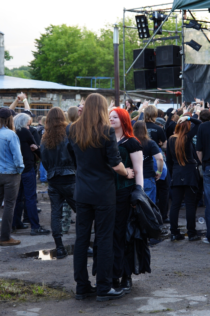 A crowd of people, mostly dressed in black, gathers outdoors for a Metalshow '09 concert. In the foreground, a person with long brown hair and a black coat faces a person with red hair in a black outfit. Stage equipment and speakers are visible in the background, and the ground is wet with puddles. Trees and buildings are also visible.