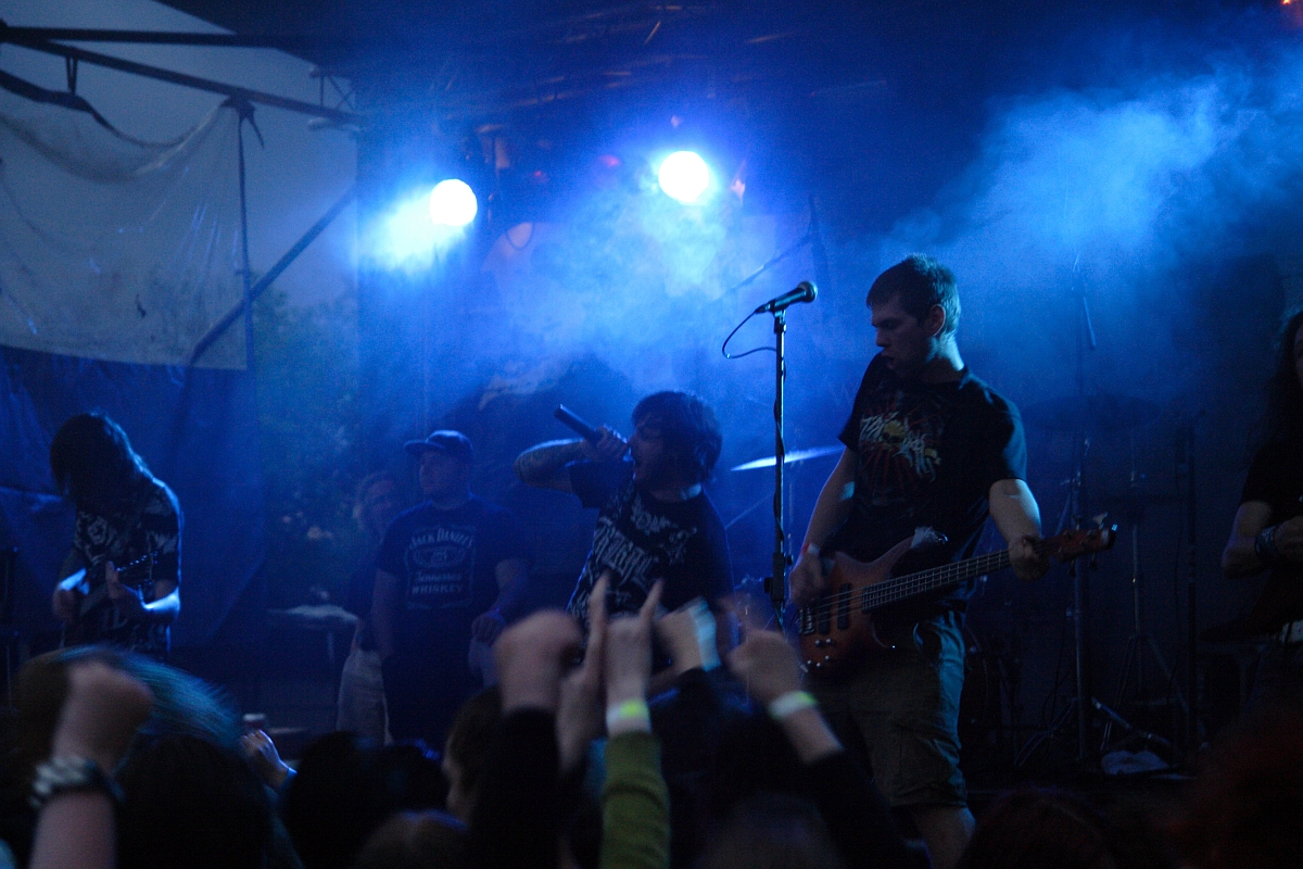At a 2009 metalshow, a rock band performs on a dimly lit stage with blue and purple lights and smoke effects. The vocalist, standing at the center, sings into a microphone while the bassist on the right plays his instrument. Fans in the foreground raise their hands, showing their excitement and appreciation.