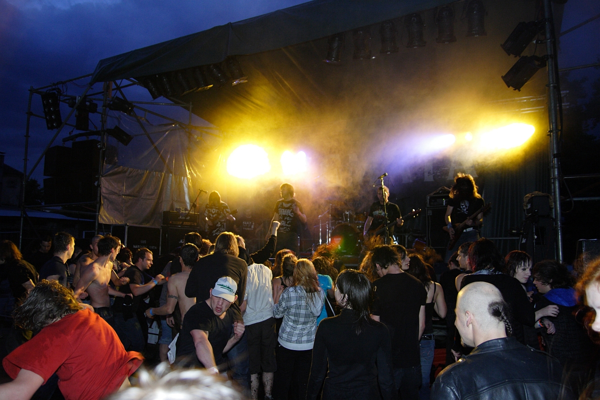 A dynamic 2009 metalshow scene at night with a rock band performing on stage under vibrant yellow and orange lights. The energetic crowd in the foreground includes fans dancing, headbanging, and raising their hands. Stage equipment and musicians are partially visible through the stage smoke.