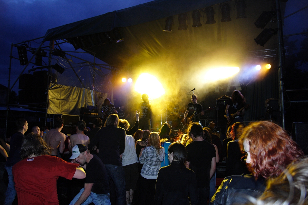 A crowd of people at an outdoor metalshow stands closely together and faces a stage where a band is performing under bright yellow and white stage lights. The atmosphere is lively, with some concertgoers visibly dancing. The sky is dark, indicating it is nighttime, reminiscent of the energetic gigs of 2009.