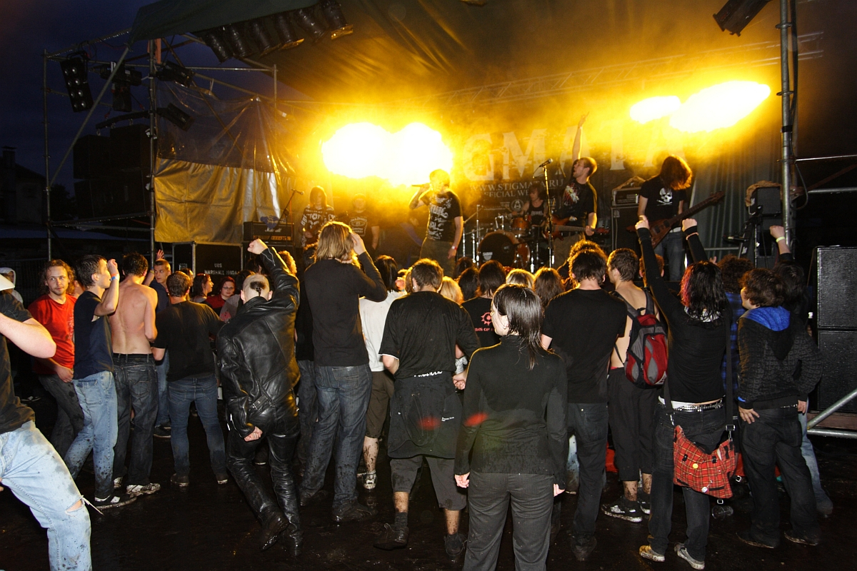 A crowd of people at an outdoor rock concert at night faces a lit-up stage. The stage, illuminated by bright yellow lights, showcases musicians performing intensely. Audience members in dark clothing headbang and raise their hands in unison. The muddy, wet ground only adds to the raw energy of Metalshow '09.