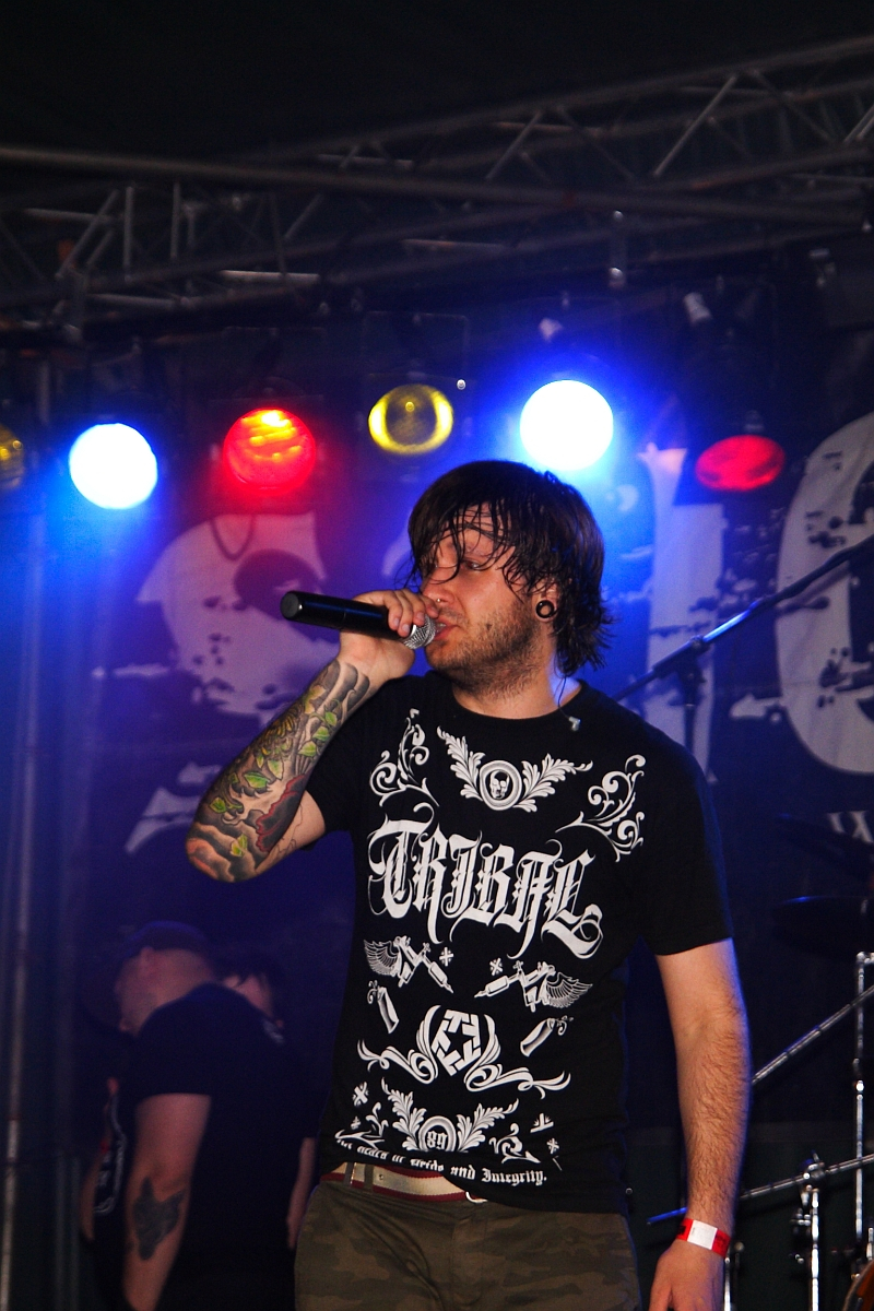 A man on stage singing into a microphone at a 2009 metalshow. He has shoulder-length dark hair, tattooed arms, and wears a black T-shirt with white ornate designs and text. In the background, colorful stage lights in red, yellow, and blue illuminate the scene, and a drummer is partially visible.