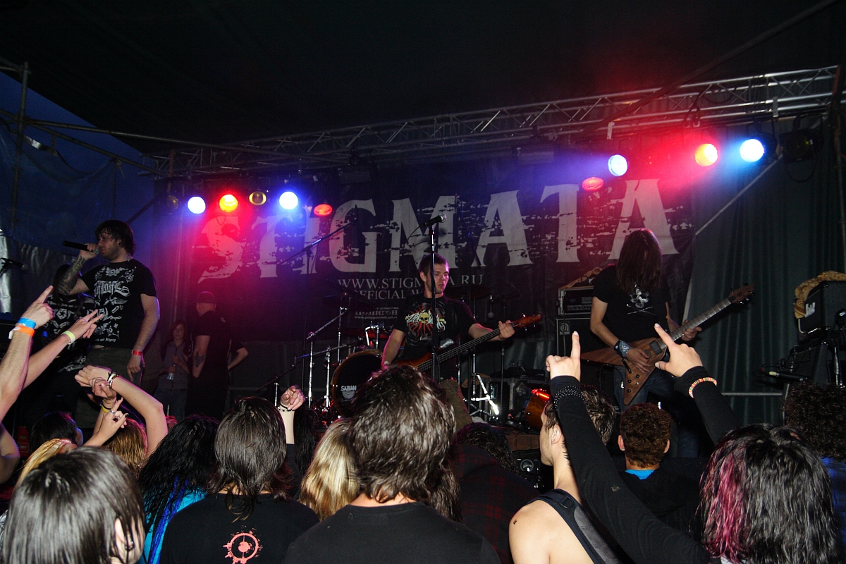 A rock band performs on a dimly lit stage with colored lights, featuring a backdrop with "STIGMATA '09" in bold letters. The lead singer holds a microphone, and the guitarist, bassist, and drummer are engaged in their instruments. The energetic crowd at this metalshow raises their hands, some forming rock-on signs.