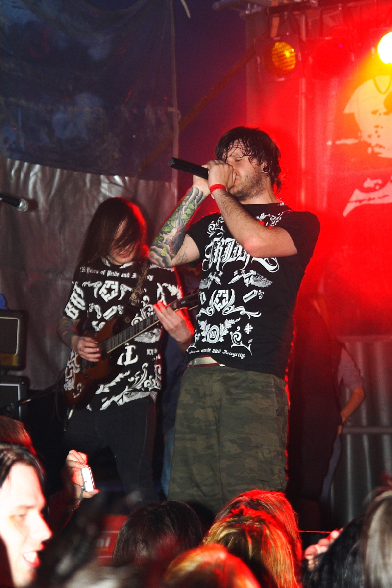 A vocalist passionately sings into a microphone while a guitarist plays beside him on stage during Metalshow '09. Both are dressed in black graphic t-shirts. The dim-lit venue is illuminated by vibrant red stage lights, and the audience, raising hands, is visible in the foreground.