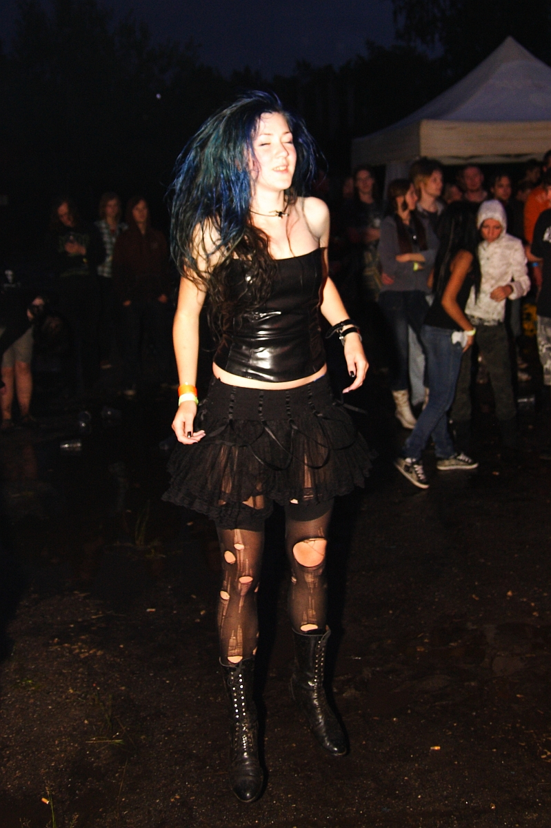 A person with blue hair is dancing outdoors at night during Metalshow 09. They are wearing a black strapless top, a black layered skirt with frayed edges, torn black tights, and black lace-up boots. Other attendees and a white tent are visible in the background. The ground appears wet and muddy.