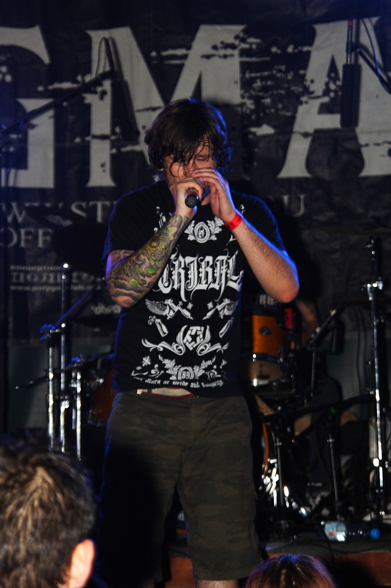 A person with long hair and tattoos on their left arm is performing on stage at a Metalshow, holding a microphone close to their mouth. They are wearing a black t-shirt with white graphics and green shorts. The background features a drum set and a dark banner with partially visible white letters.