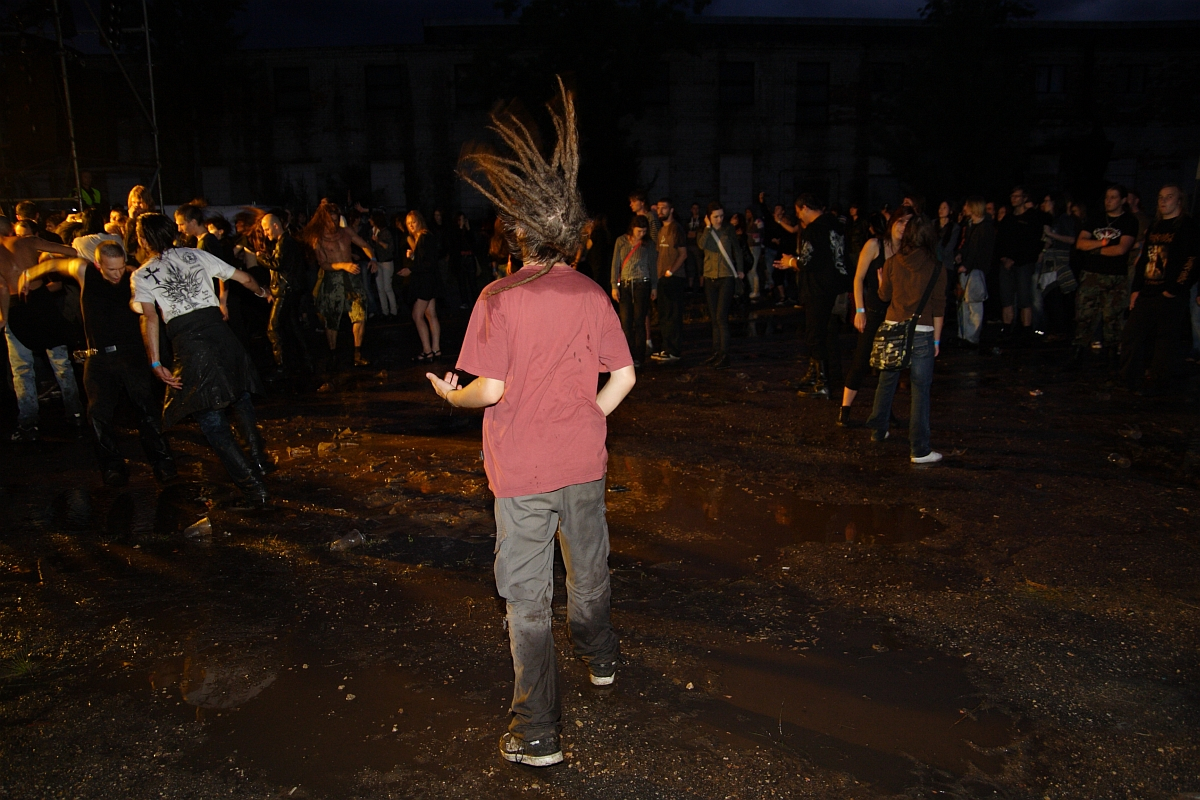 A person with dreadlocks is dancing with their back to the camera at Metalshow '09, a night-time outdoor concert. They are wearing a maroon shirt and dark pants, standing on wet, muddy ground. In the background, a crowd of people watches and mingles. The scene is dimly lit, with a dark building in the distance.