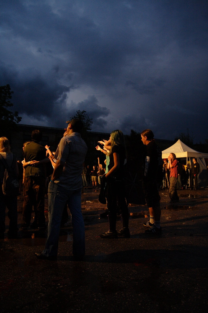 A group of people stands in a poorly lit outdoor area at dusk, facing left. The sky is dark and cloudy. Some gaze upwards, while others chat or look around. A white tent marked "Metalshow '09" is visible in the background on the right. Puddles of water are on the ground, reflecting some of the ambient light.