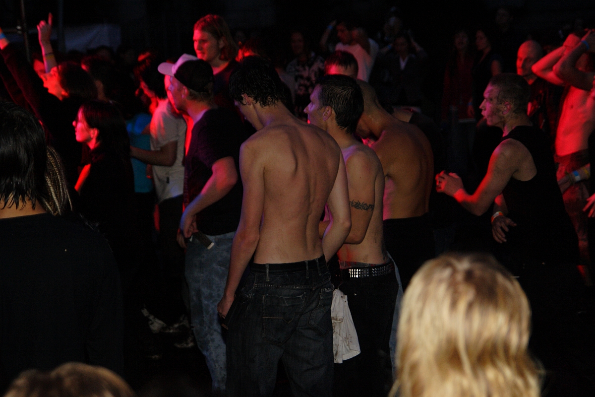 A crowded scene at Metalshow '09, with shirtless individuals enjoying the music. The audience shows a mix of energy levels, with some people raising their hands and others standing still. Colorful stage lights illuminate the crowd, casting vibrant hues of red and orange across the scene.