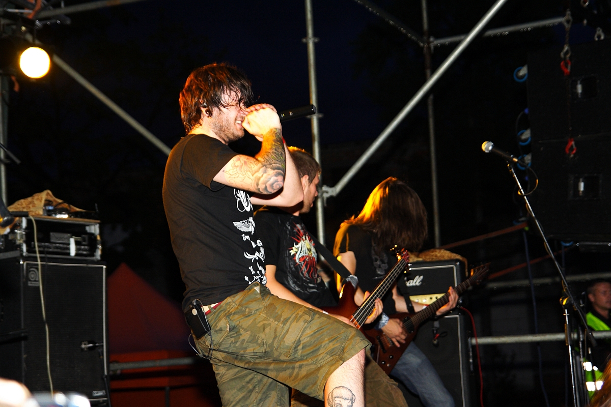 At a 2009 Metalshow, a rock band performs energetically on stage at night. The lead singer, wearing a black T-shirt with graphics and cargo shorts, passionately sings into the microphone. Two guitarists stand behind, one partially obscured. Metal scaffolding and stage lights are visible in the background.