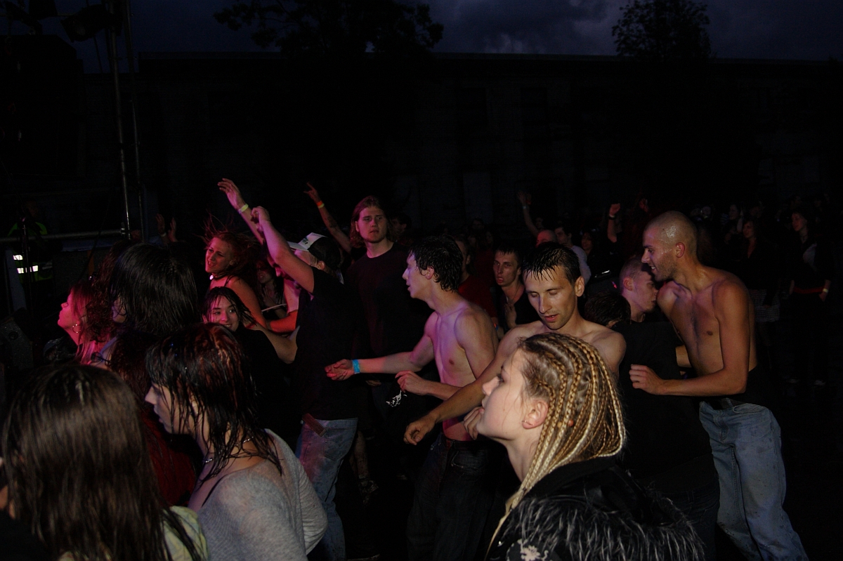 A large crowd of people is dancing and enjoying a nighttime metal show. The group is diverse, with some individuals shirtless and others wearing various clothing styles. The atmosphere appears energized, with people moving actively. Lighting from the stage casts different colors on the attendees, reminiscent of 2009 concerts.