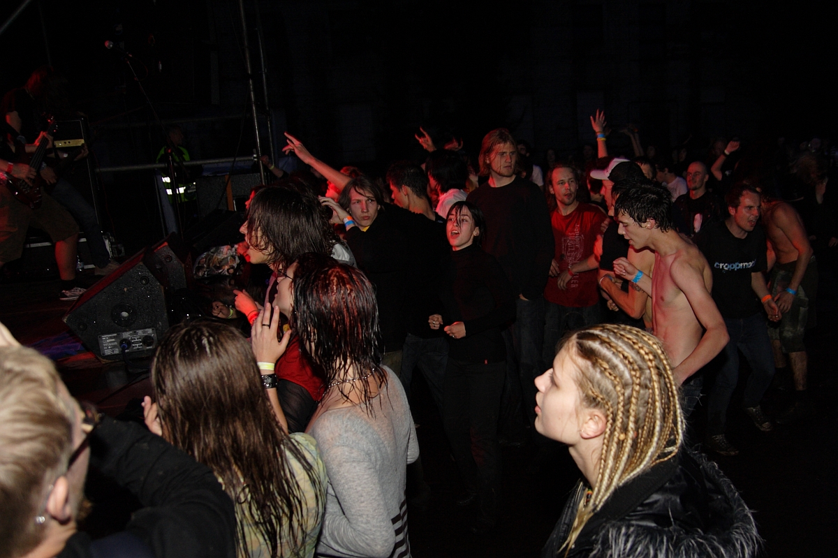 A lively crowd is attending a metalshow in a dark, indoor venue. Some are actively dancing and moving, while others are watching the performance. There is a mix of casual and more expressive attire, including one individual with braided hair in the foreground. This event has all the energy of 2009.